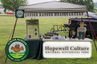 A table under a tent, outdoors with a cardboard cutout of a junior ranger child and junior ranger materials on top of tables