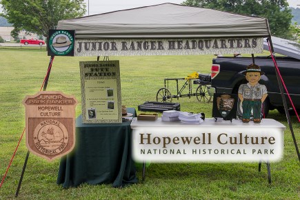 A table under a tent, outdoors with a cardboard cutout of a junior ranger child and junior ranger materials on top of tables