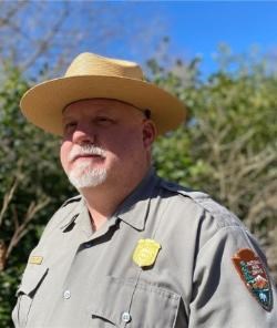 A man wearing a tan flat hat and gray shirt with arrowhead logo on the sleeve under a blue sky