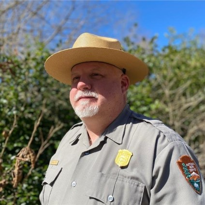 Alford Headshot A man wearing a tan flat hat and a gray ranger shirt stares into the distance under a blue sky
