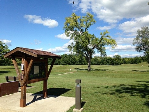 A wooden informational sign in the foreground and several grass-covered mounds in the background, under a blue sky.