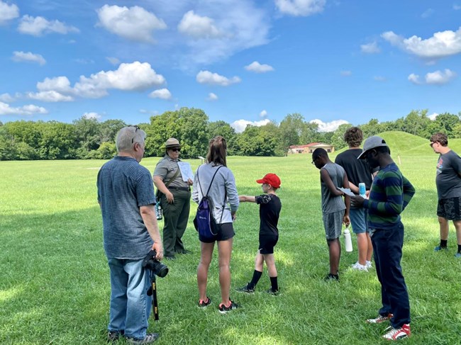 a group of people stand near a park ranger in uniform leading a talk in a sunny field.