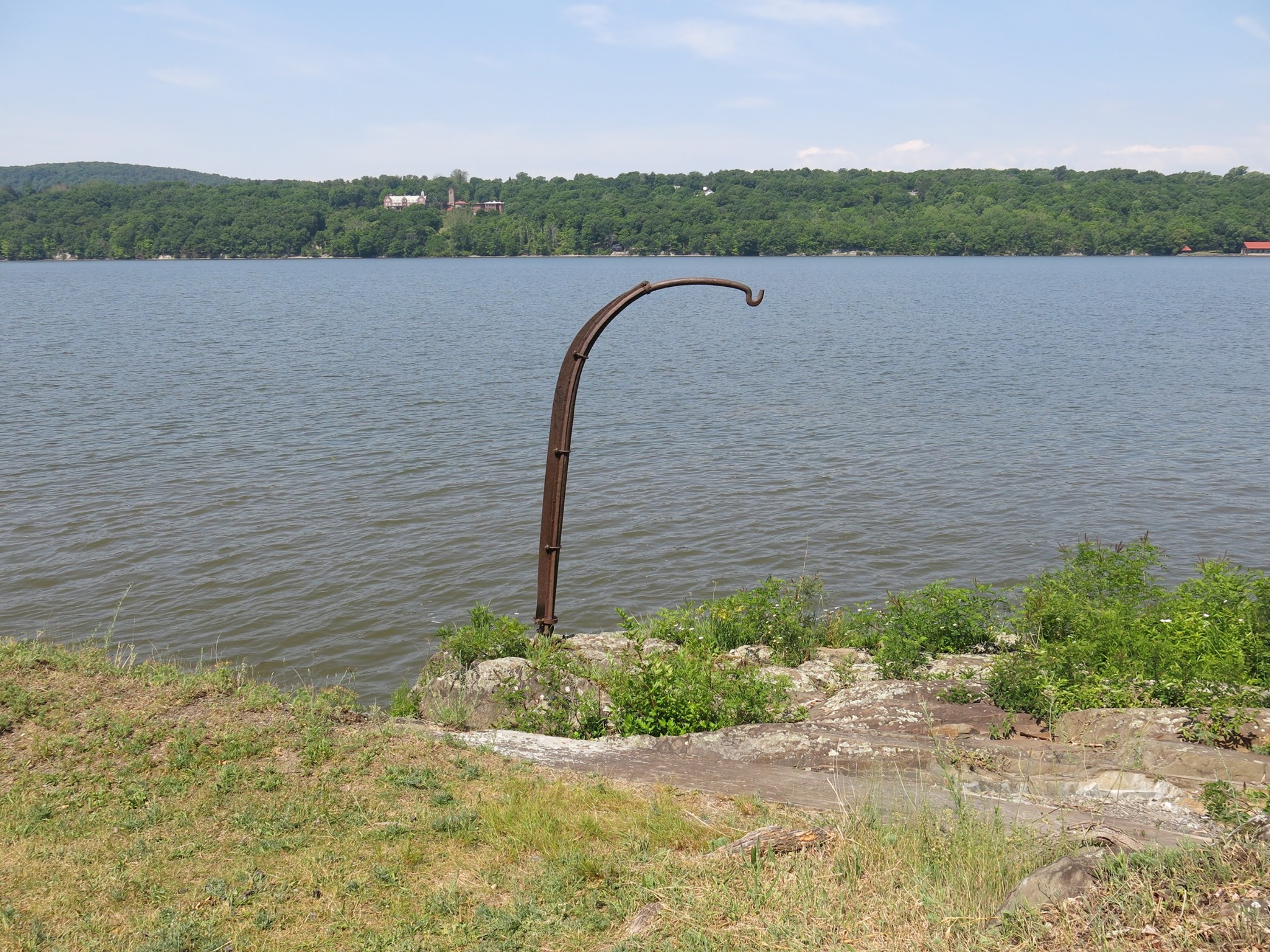 A metal hook stands on a rocky shoreline of a river. Buildings are visible across the river.