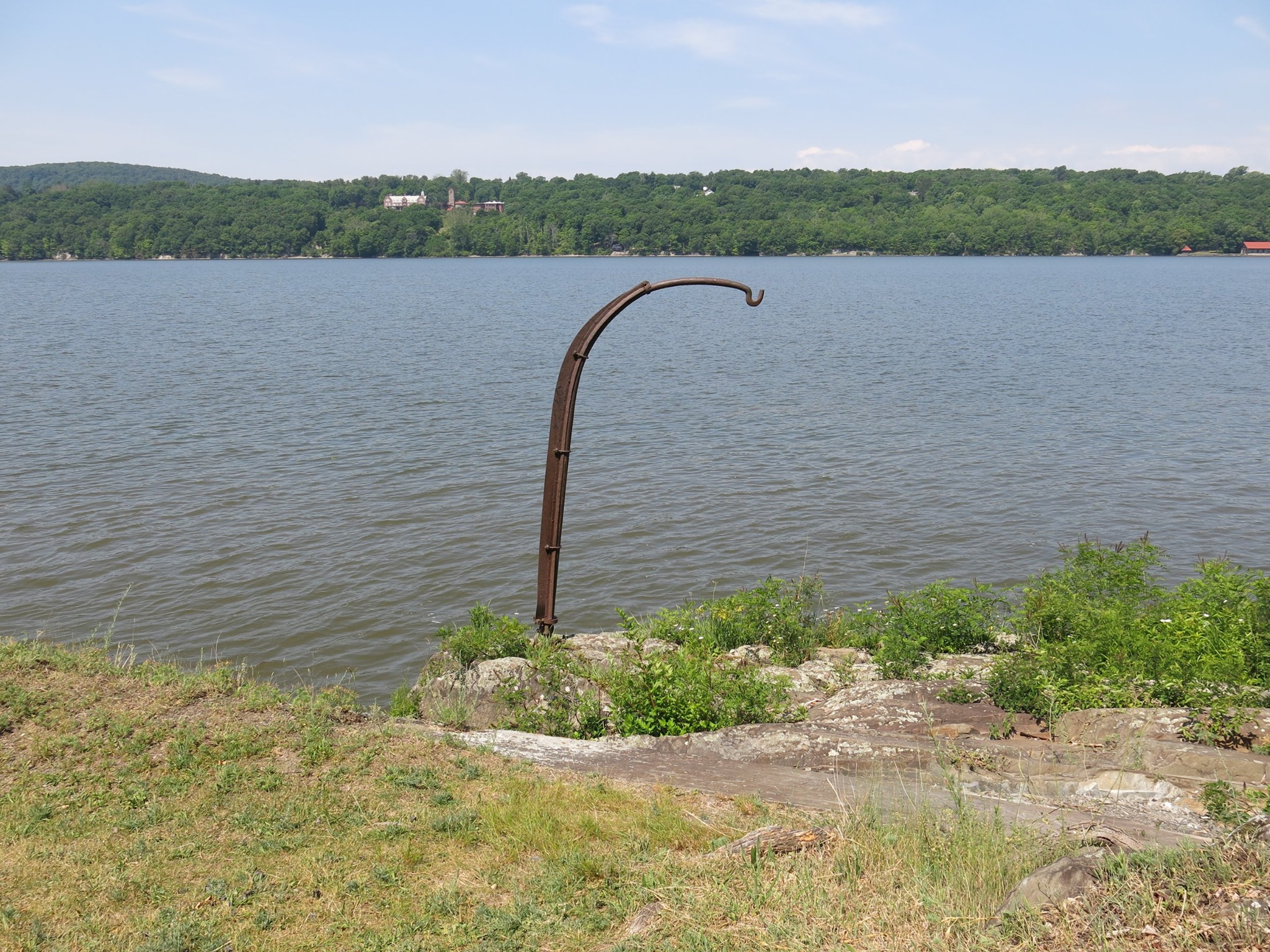 The boat hook at Bard Rock A metal hook stands on a rocky shoreline of a river. Buildings are visible across the river.