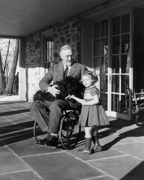 FDR seated in a wheelchair with a dog in his lap and a young girl standing next to him.