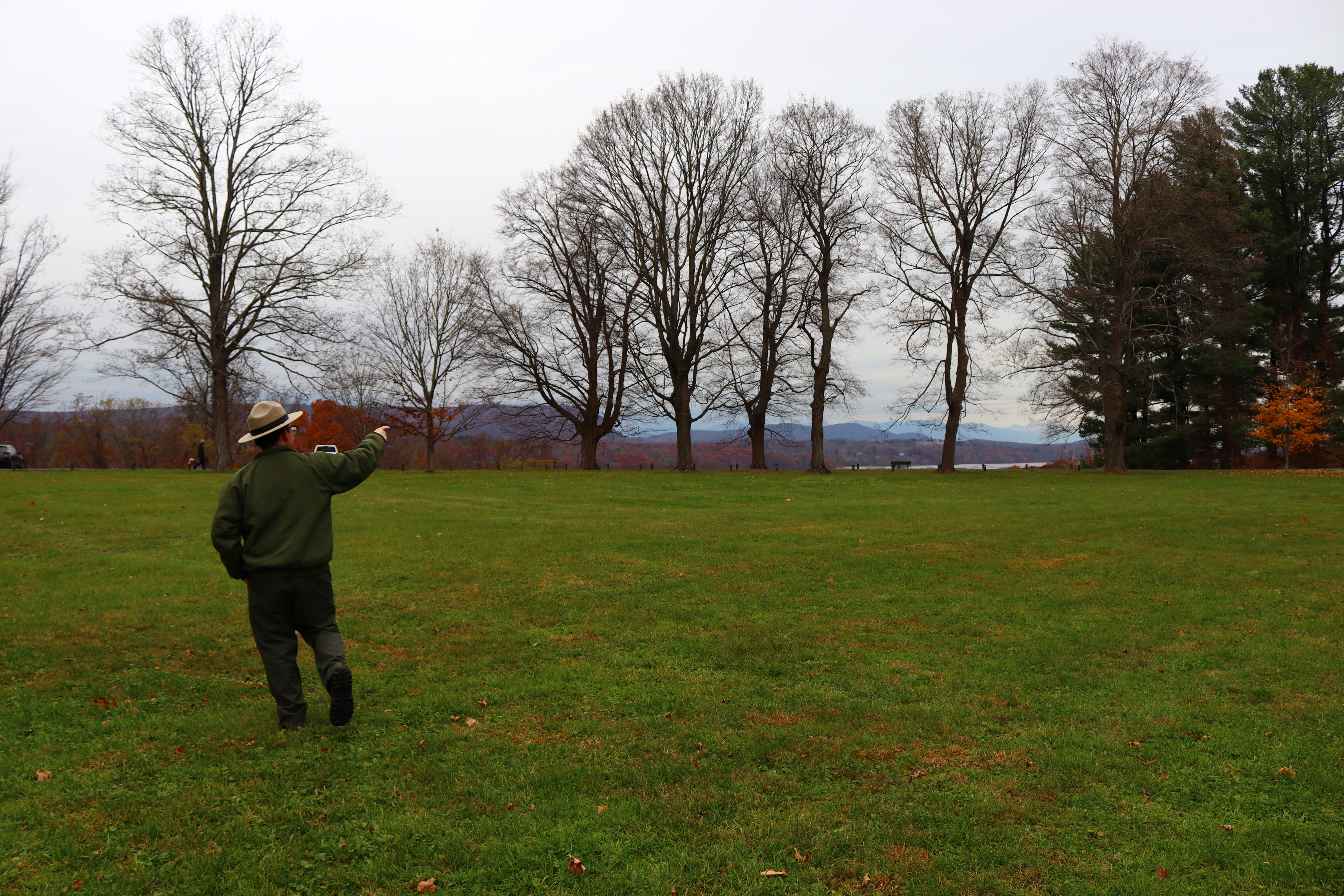 A male park ranger points to his right as he walks across a grassy field. Mountains and a river are visible in the background.