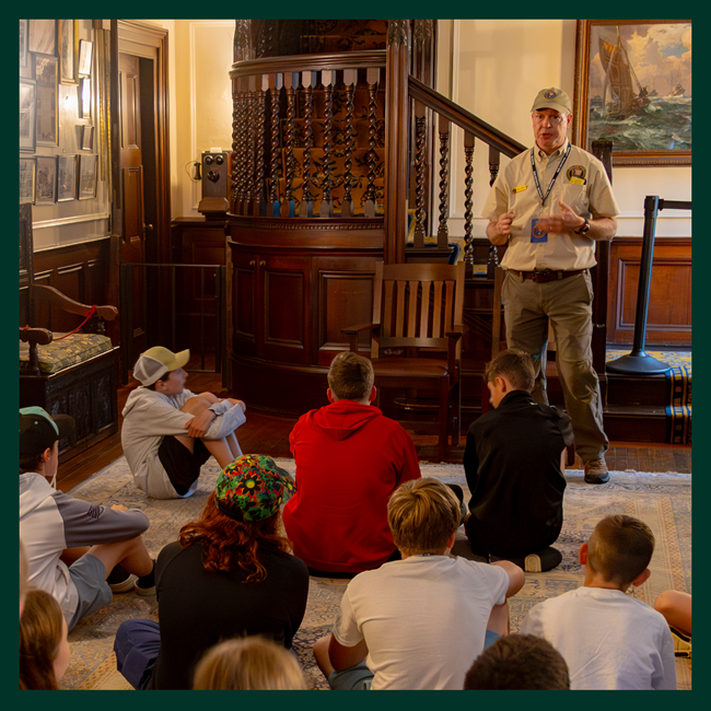A photo of a man in NPS volunteer clothing standing inside an historic home. He speaks to children who are listening while sitting on a rug in the home's foyer.