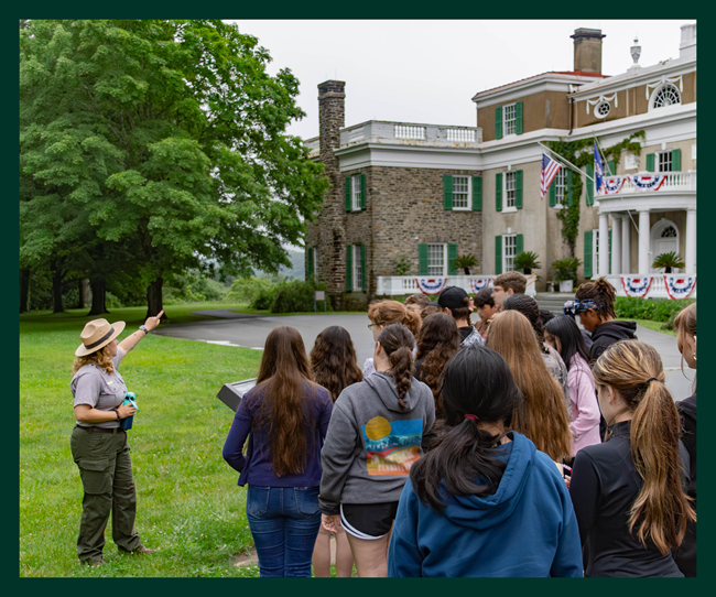 A park ranger points to the Home of Franklin Roosevelt as a group of middle school children gathers around the ranger.
