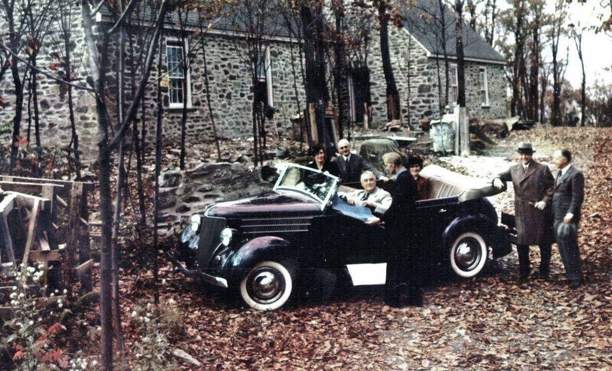 A man (FDR) seated in a car surrounded by several people at a house under consturction.
