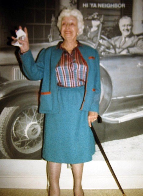 An elderly Toi Bachelder stands with a cane in front of a large image of FDR driving an automobile.