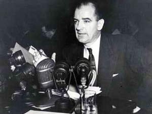 A man (Joseph McCarthy) is seated and speaking into a group of microphones.