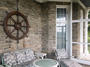 A screened porch with large ship's wheel hanging above upholstered furniture.