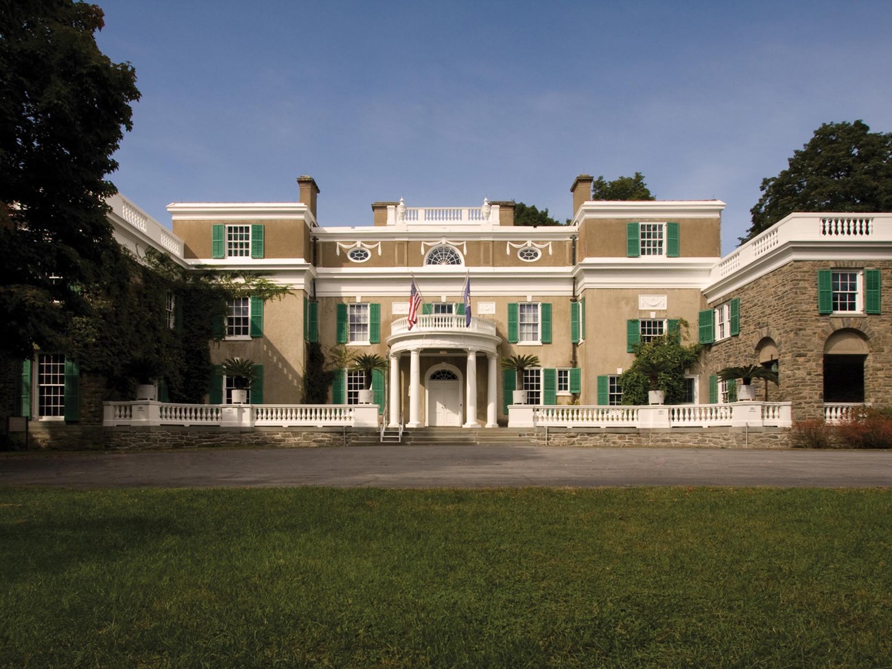 A two-story stone and stucco house with central portico on a lush lawn surrounded by trees.