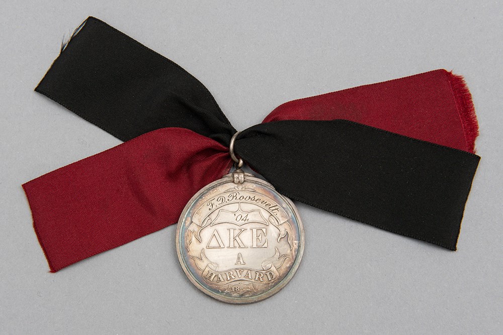 A silver medal with insignia of the Greek letters DKE and engraved "F. D. Roosevelt" and "04," attached to a blue and crimson ribbon.