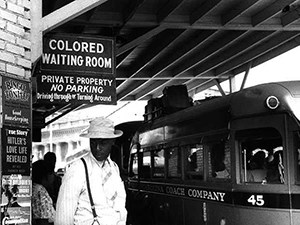 A man of African descent stands outside a bus station with sign mounted to building stating "Colored Waiting Room."