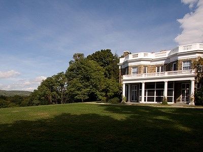 A house of stone with white painted columns, balustrade roof, and screened porch on a green lawn.