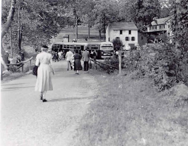 People walking toward a buses that is parked near the furnace stack. Office/Store and Ironmaster's Mansion in view.