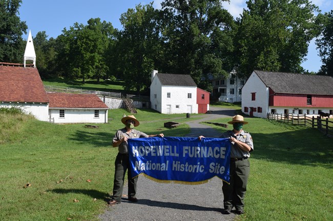 Two Park Rangers with masks pose for a picture with a Hopewell Furnace Banner. Cast House, Office/Store, Barn and Ironmaster's Mansion in background.