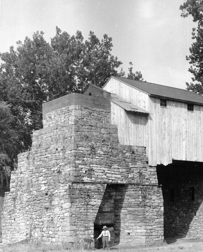 A man (perhaps a Park Ranger) stands in front of the furnace stack. Bridge House in view.