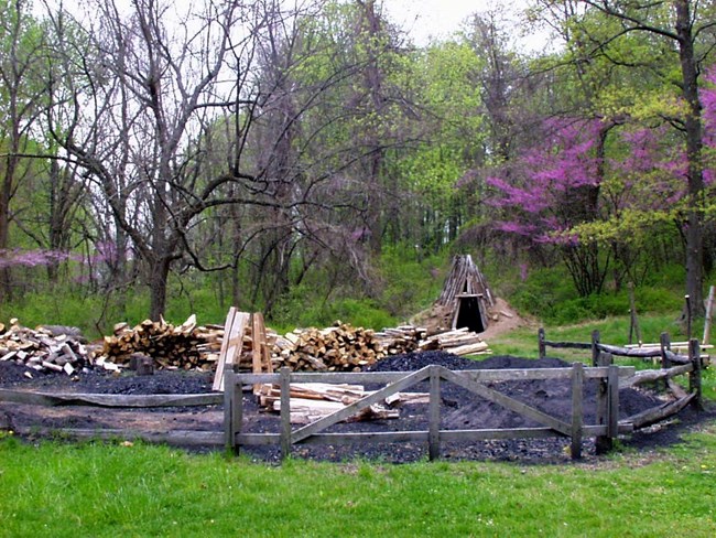 Pit with charcoal and wood scattered around the area with a hut made of earth and wood built in background.