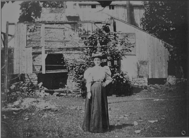 Black and white photograph of a woman standing in front of the furnace stack. Debris from Cast House and related buildings is scattered on the ground behind her. The photograph appears to be from the late-1800s or early-1900s.