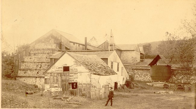 View of deteriorating Cast House with a man standing in foreground. Back of furnace stack is also in view.