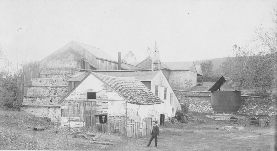 Black and white image of man standing in front of wooden building with triangular roof. Exposed conical brick structure stands to the left of the wooden building.