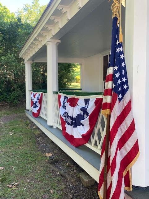 NPS Photo An American flag sits in front of the Ironmaster's Mansion porch. The porch bannisters are covered in American flag decorative bunting.