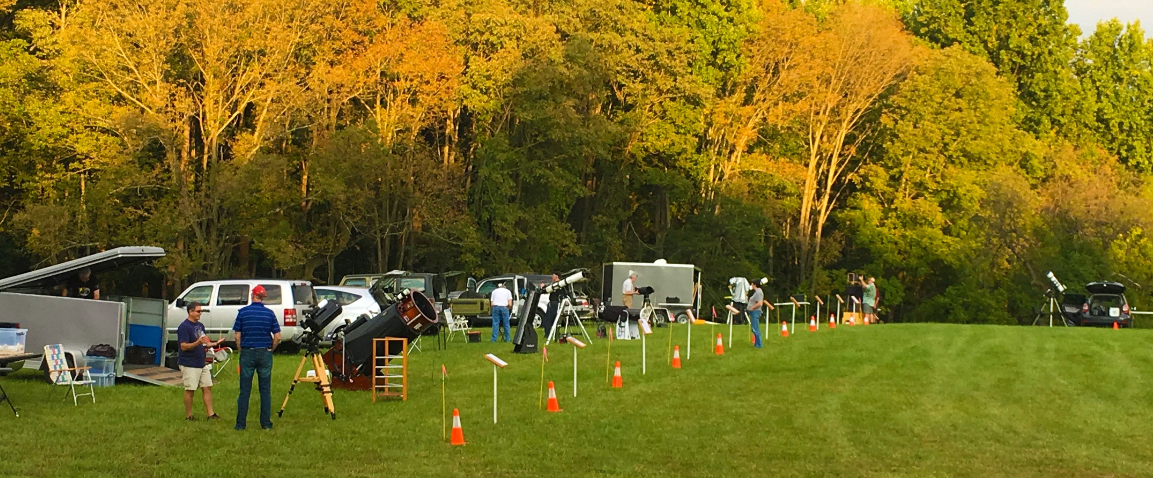 Starfest People stand in a grassy field at sunset with telescpes and traffic cones set up in a long line.