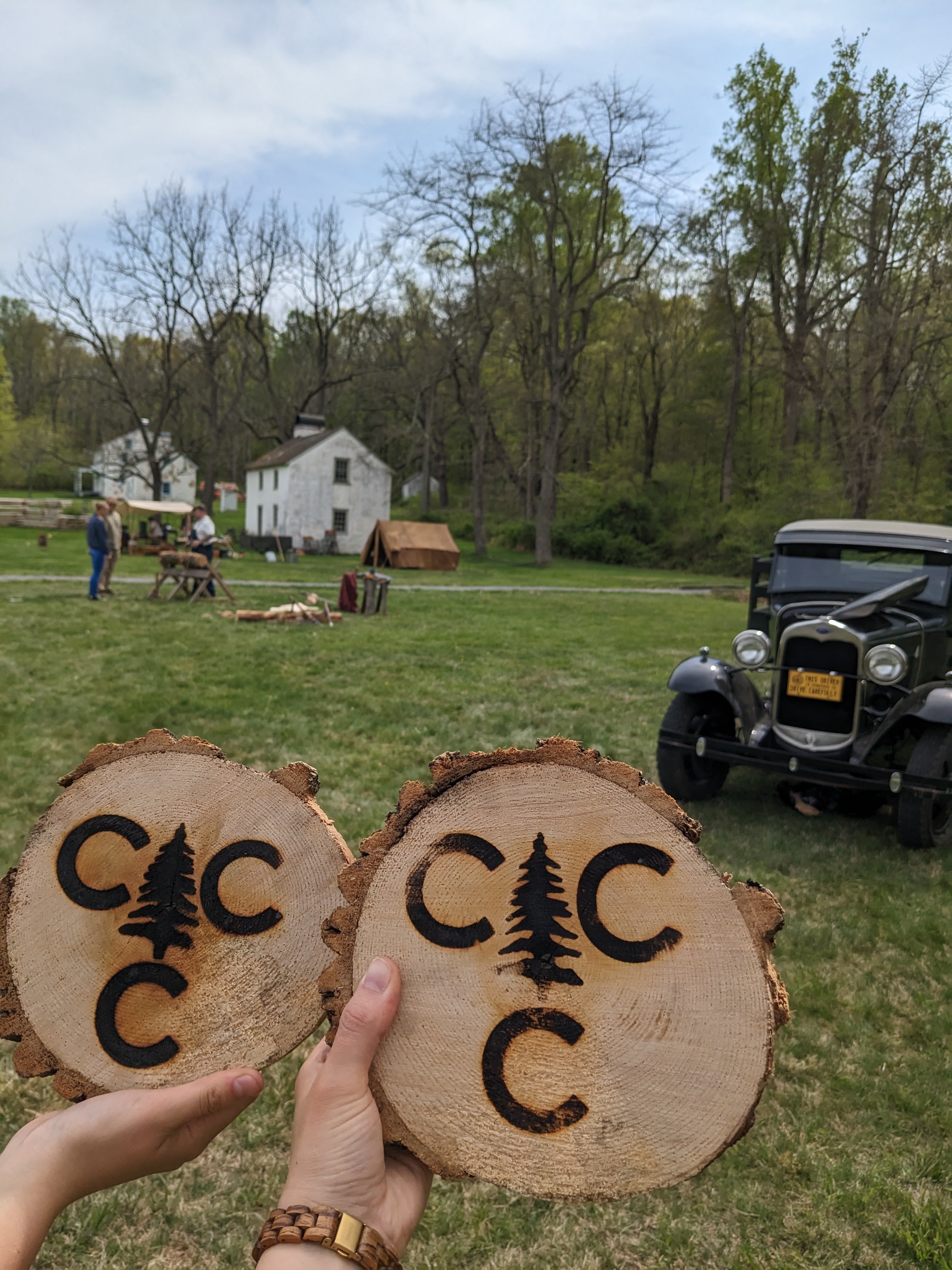 NPS Photo/ CCC Encampment two people holding wood pieces that are branded with the CCC logo. In the background is a 1930s era truck, some tents and people walking through the historic site.