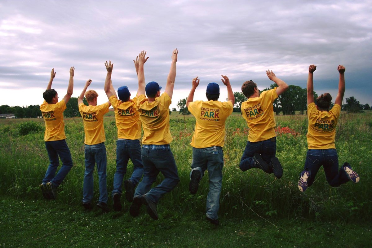High School students enjoy volunteering at Homestead National Monument. Seven high school students jump in the air with their hands up and with their back to the camera. They face a prairie and an overcast sky.