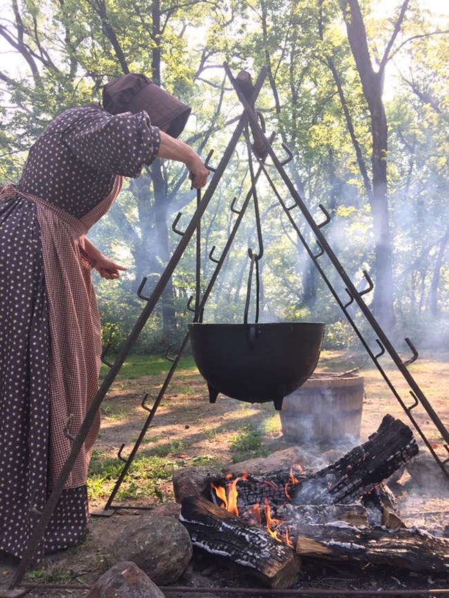 A woman in a prairie dress and bonnet stands next to large black pot hanging over a fire.