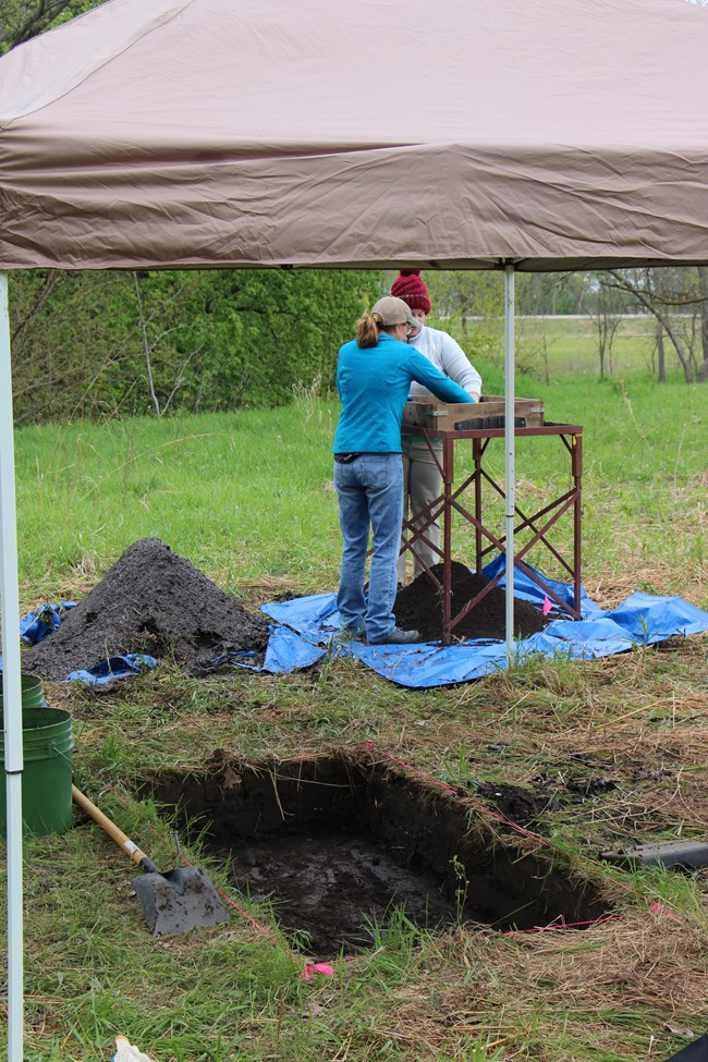 Two people sifting dirt next to an excavation pit