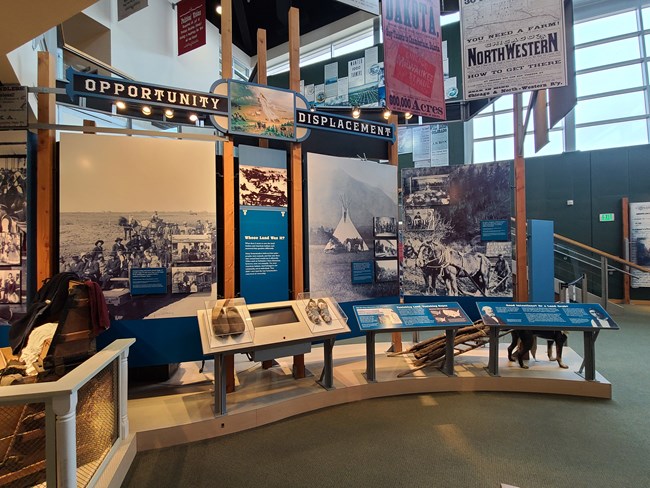 Museum exhibit with text panels and historic images about American Indians. To the left is a trunk full of clothing. In the center are two pairs of shoes. On the right is a model of a dog pulling a travois.