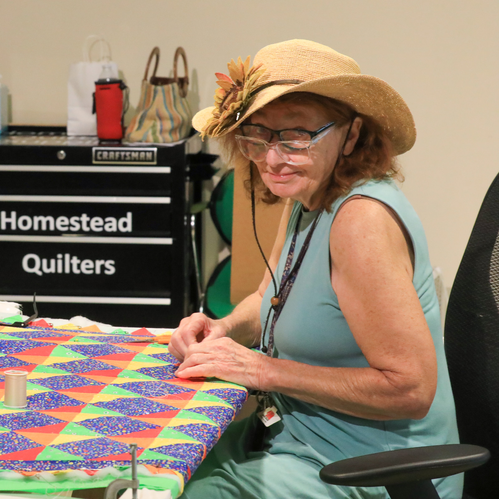 AIR Anita Bauman Woman with red hair under straw hat looks at camera through 2 sets of glasses while sewing.