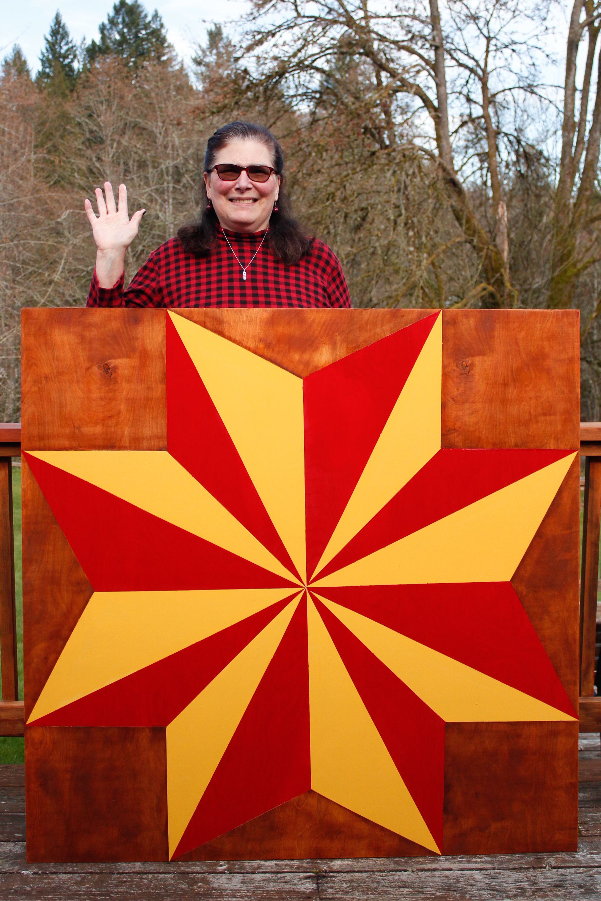Vickie MacMillan A woman stands behind a large wooden board painted in a quilt square pattern. She waves.