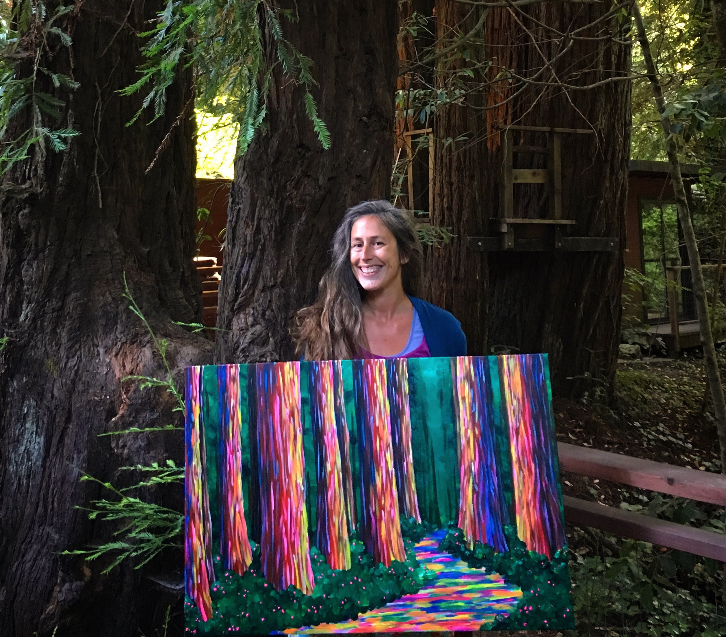 A woman stands among large trees holding a painting of colorful trees and a path.