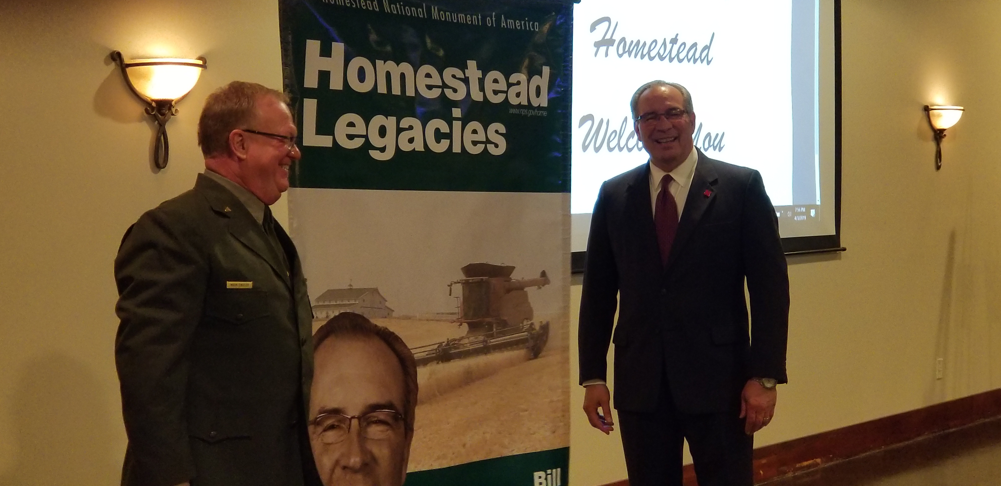 Homestead National Monument of America Superintendent Mark Engler with Bill Moos at the Homestead Legacy Banner unveiling. A smiling man in park service uniform looks across a banner at another man in a suit.