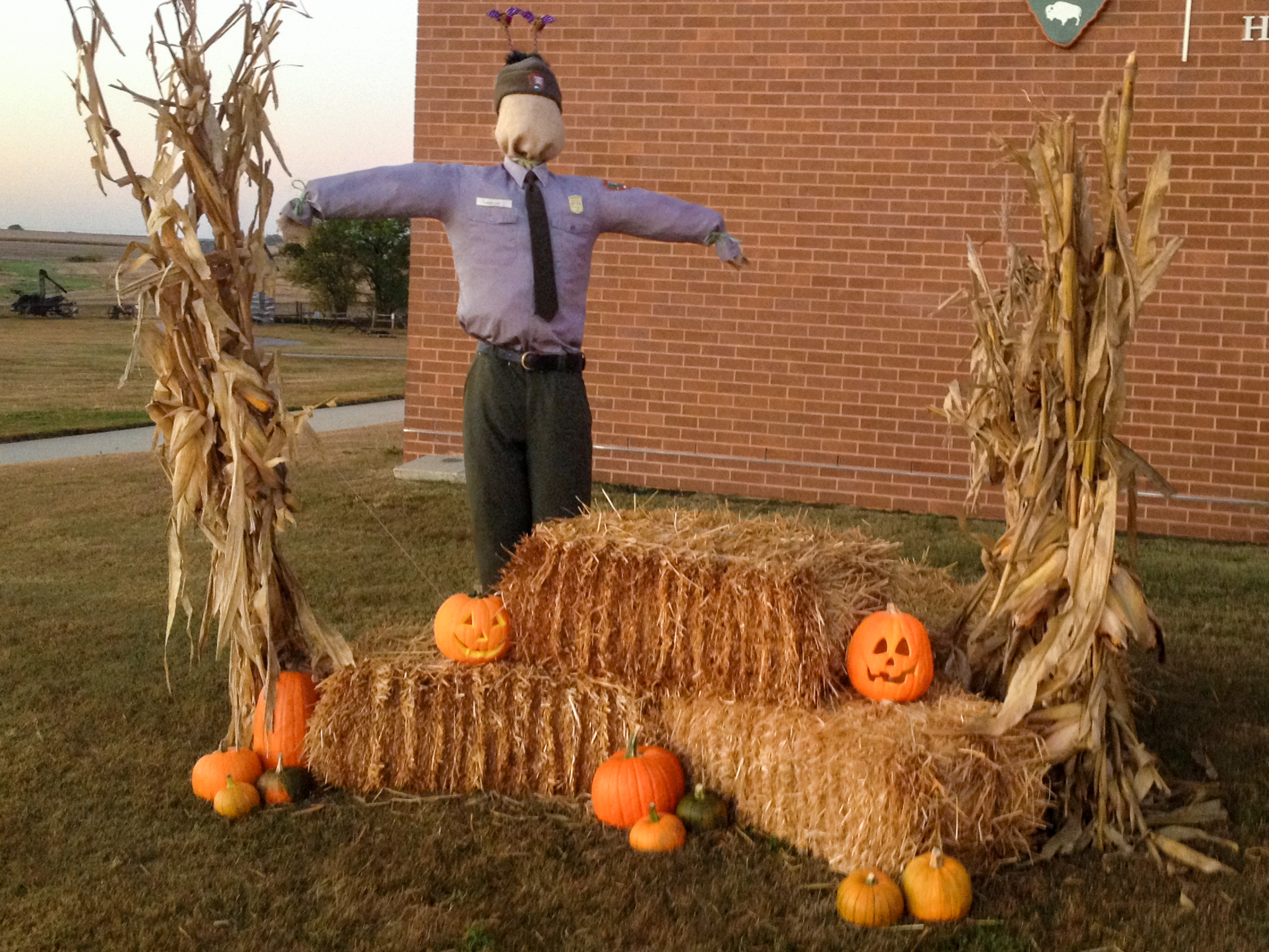 Come join us on October 26, 2024, at 4pm for Howling Homestead! Hay bales, corn stalks, pumpkins, and a uniformed NPS scarecrow in front of a brick building.