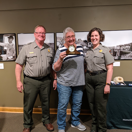 2019 VIP of the Year A woman stands holding a clock with an engraved plate on it. A uniformed park ranger stands on either side of her.