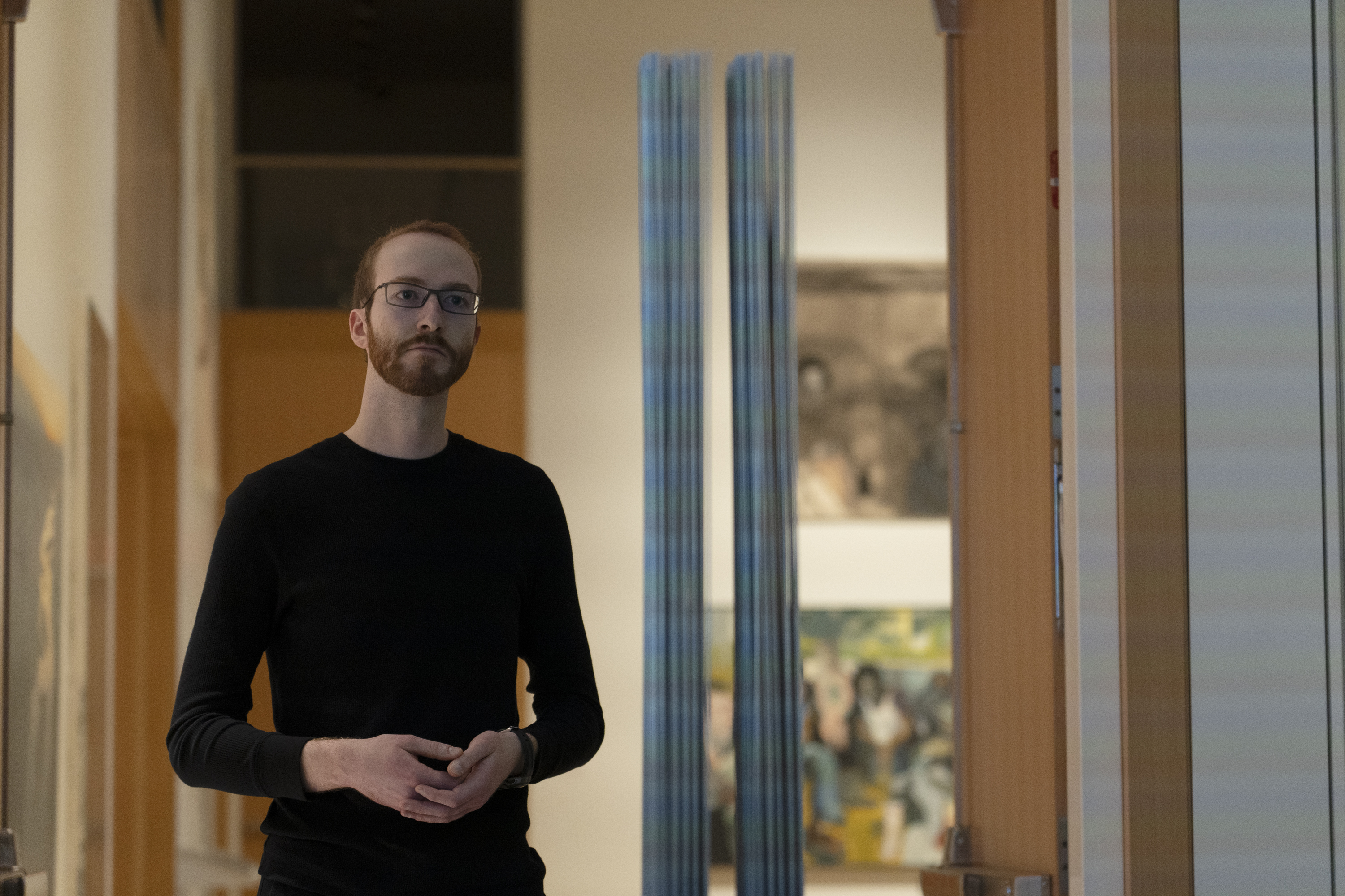 Dr. Benjamin Justis A man wearing glasses and a longsleeve black shirt stands with his hands in front of him, fingertips touching.