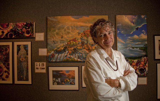 Nancy Lehenbauer Marshall A woman stands with her arms crossed in front of a wall displaying multiple paintings.