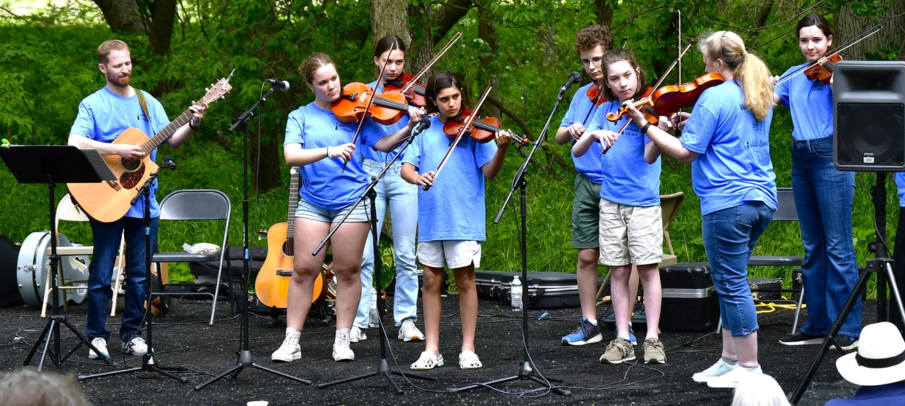 Seven fiddlers and a guitarist play on an outdoor stage in front of a crowd.