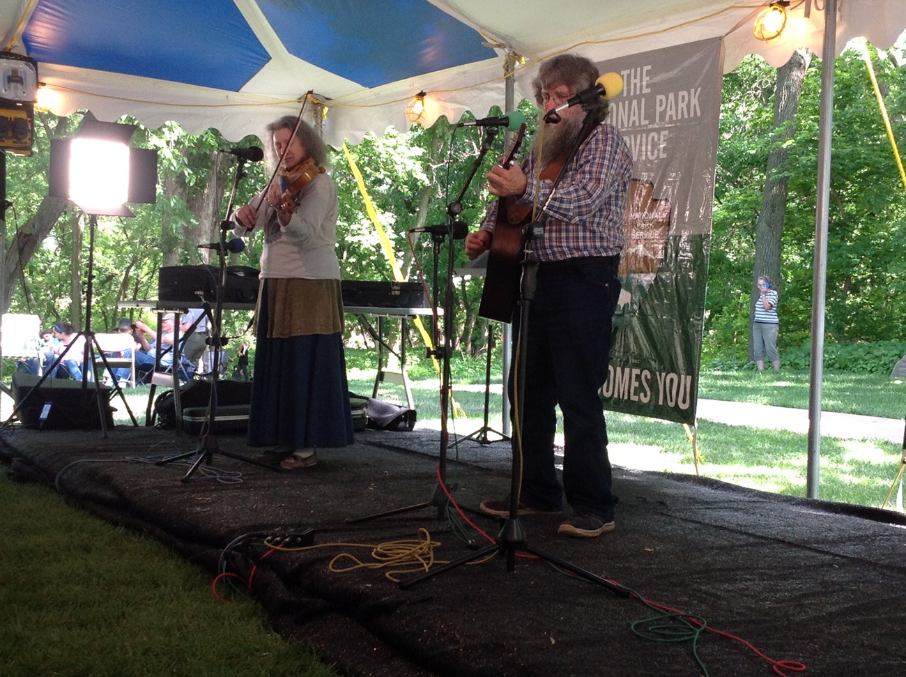 A woman with a fiddle stands on a stage next to a man playing the guitar.