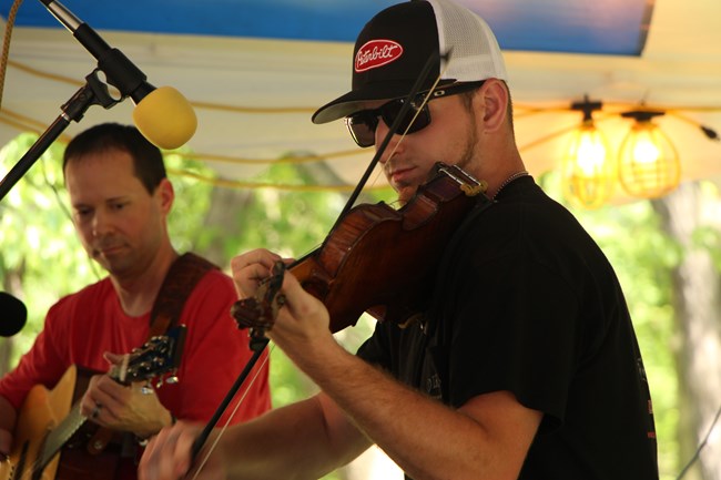 A man wearing sunglasses and a ballcap plays the fiddle while a man in the background plays a guitar