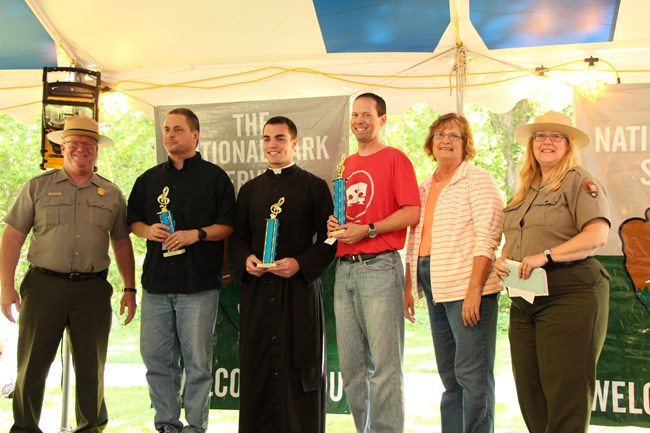 Two park rangers stand on either side of four people, three of whom are holding trophies.