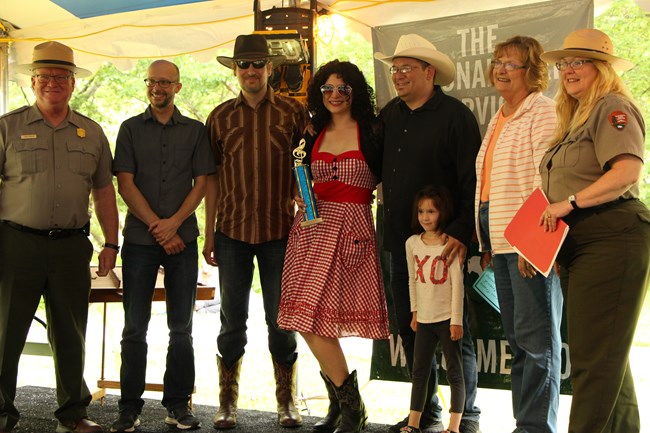 Two park rangers stand on either side of a group of five people. One of whom holds a trophy.