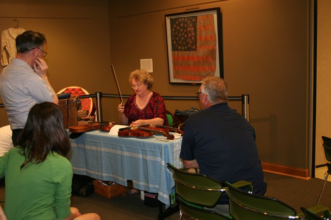 A woman is seated at a table holding a fiddle and bow while a group of people stand looking at her.