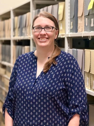A woman in a blue shirt wearing glasses is smiling in front of shelving