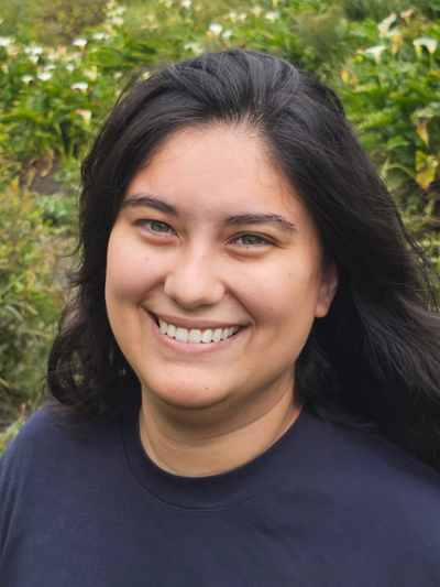 A woman with long, brown hair in a blue shirt is smiling. Behind her is green foliage.